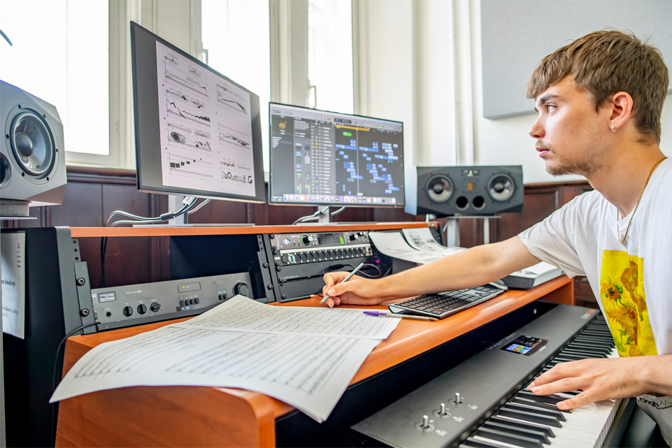 A male student looking at a computer monitor with music software on it, with technical equipment used to compose music.